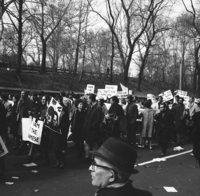 Demonstration against Vietnam War: Union Groups, Shoot ID: 409, Negative: 21. Image of a black and white photograph. In the photograph, a crowd of people stand in the middle of a street holding posters. Behind the crowd, there is a hill with bare trees. 