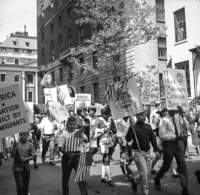 Labor Day (Other Street Scenes), Shoot ID: 410, Negative 12. Image of a black and white photograph. In the photograph, a group of people carrying posters and US flags walk down a street. In the background, there are several tall buildings. The photograph includes posters that say "I Like Garments With This Union Label", "Equal Opportunity", "Educated Children Today Mean a Stronger Nation Tomorrow", and "Union Made ILGWU AFL-CIO Int. Ladies Garment Workers Union" (parts of the posters' text are covered or unclear). Full text includes "I Like Garments With This Union Label Equal Opportunity Educated Children Today Mean a Stronger Nation Tomorrow Union Made ILGWU AFL-CIO Int. Ladies Garment Workers Union".