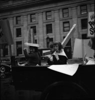 May Day, Shoot ID: 256, Negative: 34. Image of a black and white photograph. In the photograph, three girls ride in the back of a truck with posters. In the background, there is a building. 