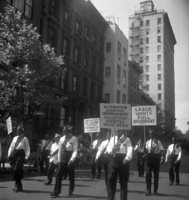 Labor Day, Slogans include "Pass Anti-Scab Legislation" and "TWU Supports Freedom Riders", Shoot ID: 416, Negative 34. Image of a black and white photograph. In the photograph, a group of men in matching uniforms walks down a street with posters. The posters say "Labor Wants Full Employment", "Automation Causing Unemployment Solution: Shorter Work Week", and "Register So You Can Vote For Friends of Labor". A crowd watches from behind a police line on the sidewalk. In the background, there are several buildings. Full text includes "Labor Wants Full Employment Automation Causing Unemployment Solution: Shorter Work Week Register So You Can Vote For Friends of Labor".