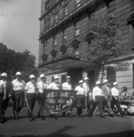Labor Day, Shoot ID: 406, Negative 26. Image of a black and white photograph. In the photograph, a line of men in hard hats walk down a street with a banner. The banner says "Safety Committee Local Union No. 3" (part of the banner is covered and unclear). A few people watch from behind a police line on the sidewalk. Behind the spectators, there is a large building. In the background, there are several trees. Full text includes "Safety Committee Local Union No. 3".