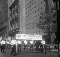 Labor Day, Shoot ID: 406, Negative 8. Image of a black and white photograph. In the photograph, a group of men in matching uniforms walk down a street with posters. In front of the group, two men carry a large banner that says "Cutters' Union Local 10 ILGWU". The group walks by several tall buildings. Full text includes "Cutters' Union Local 10 ILGWU". 