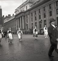 May Day, Shoot ID: 256, Negative: 42. Image of a black and white photograph. In the photograph, a small group of women in dresses walks down a street. On the far side of the street, there is a large building and pedestrians walking along the side of it. In the background, there are several policemen, buildings, and cars. 