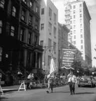 Labor Day, Slogans include "Pass Anti-Scab Legislation" and "TWU Supports Freedom Riders", Shoot ID: 416, Negative 28. Image of a black and white photograph. In the photograph, there are two men with flags walking down a street. One man carries a US flag and the other carries a flag or banner that says "Theatrical Stage Employees Local No. 4 Brooklyn. N.Y. LA.T.S.E. & M.P.M.O. of United States & Canada Affiliated with A.F. of L. Since 1888" (part of the banner's text is covered and unclear). Behind the men, there is a car driving down the street. A crowd watches from behind a police line on the sidewalk. In the background, there are several buildings. Full text includes "Theatrical Stage Employees Local No. 4 Brooklyn. N.Y. LA.T.S.E. & M.P.M.O. of United States & Canada Affiliated with A.F. of L. Since 1888".