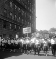 Labor Day, Shoot ID: 417, Negative 9. Image of a black and white photograph. In the photograph, a group of people in matching graduation caps walks down a street with posters. The group walks next to a large building. In the background, there are trees. The photograph includes posters that say "Scholarship Winners", "1955 27 Scholarships", and "1950 3 Scholarships". Full text includes "Scholarship Winners 1955 27 Scholarships 1950 3 Scholarships".
