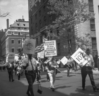 Labor Day (Other Street Scenes), Shoot ID: 410, Negative 19. Image of a black and white photograph. In the photograph, a crowd of people with posters and US flags walk down a street. In the background, there are several tall buildings. The photograph includes posters that say "Local 102 ILGWU", "Educated Children Today Mean a Stronger Nation Tomorrow", "End Poverty", and "Union Made ILGWU Int. Ladies Garment Workers Union" (parts of the posters' text are covered or unclear). Full text includes "Local 102 ILGWU Educated Children Today Mean a Stronger Nation Tomorrow End Poverty Union Made ILGWU Int. Ladies Garment Workers Union".
