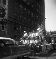 Labor Day, Shoot ID: 417, Negative 3. Image of a black and white photograph. In the photograph, a group of men sit on a float being pulled by a car. The float includes banners that say "Local 3", "Local 3 - IBEW", and "New York City Labor Advocates 1964 World's Fair" (parts of the banners' text are covered or unclear). A bus follows the float. A crowd watches from the sidewalk. In the background, there is a large building. Full text includes "Local 3 Local 3 - IBEW New York City Labor Advocates 1964 World's Fair".