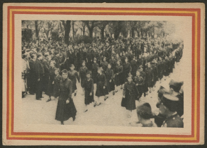 Front of postcard (recto). Image of a photograph of girls in uniform marching down a street. In the background, there is a crowd saluting the girls with right arms extended from the neck into the air with a straightened hand. Behind the crowd, there is a line of trees. In the lower right corner, there is part of another group of spectators. The photograph has a red and yellow striped border. 