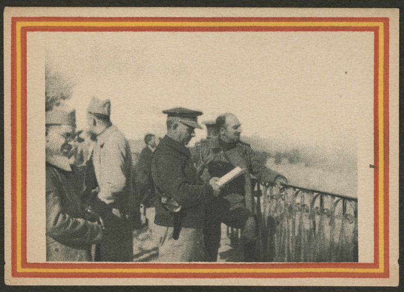 Front of postcard (recto). Image of a photograph of a group of men in uniform standing on a railed ledge. One man looks down at a piece of paper. The photograph has a red and yellow striped border.  